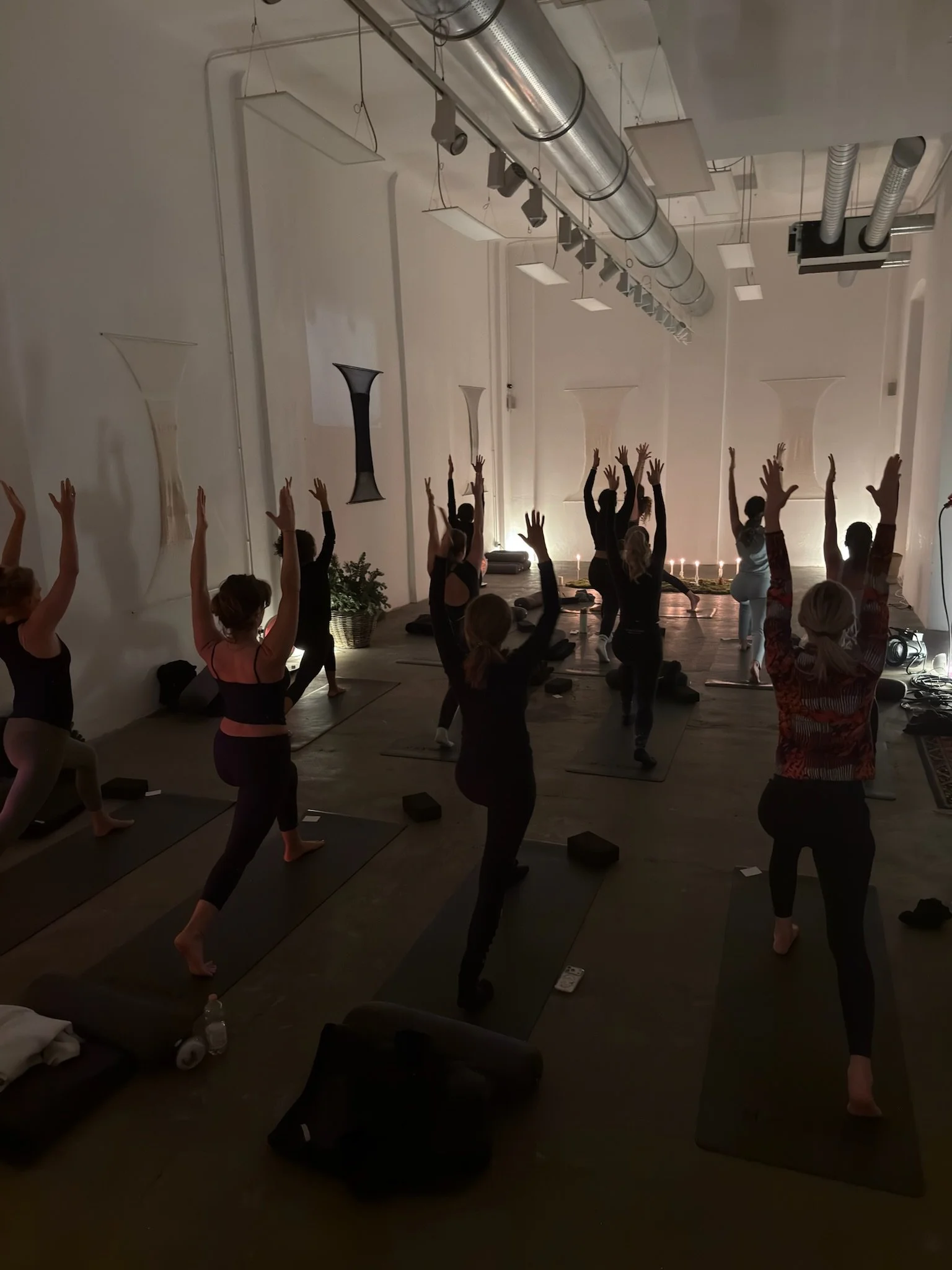 A group of people participating in a yoga class in a dimly lit room, lifting their arms upward while standing on yoga mats.