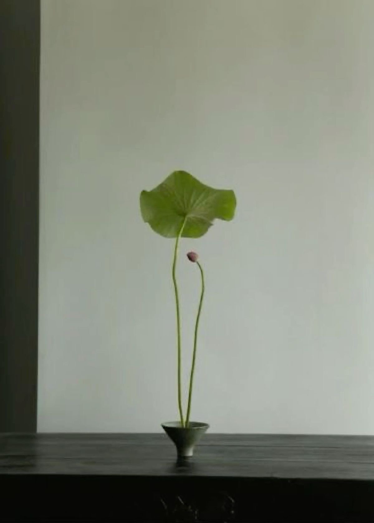 A leaf and a flower in a small black vase placed on a wooden surface against a plain light-colored wall.