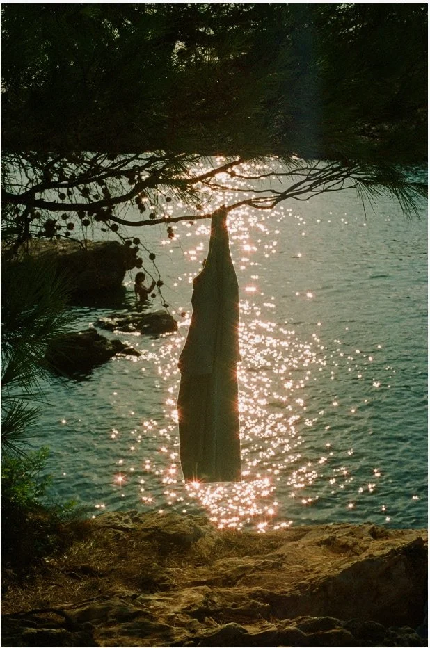 A scenic view of a lakeside with sunlight reflecting off the water. A piece of clothing, likely a towel or shirt, hangs from a tree branch over the water. In the background, a person is swimming near rocks.