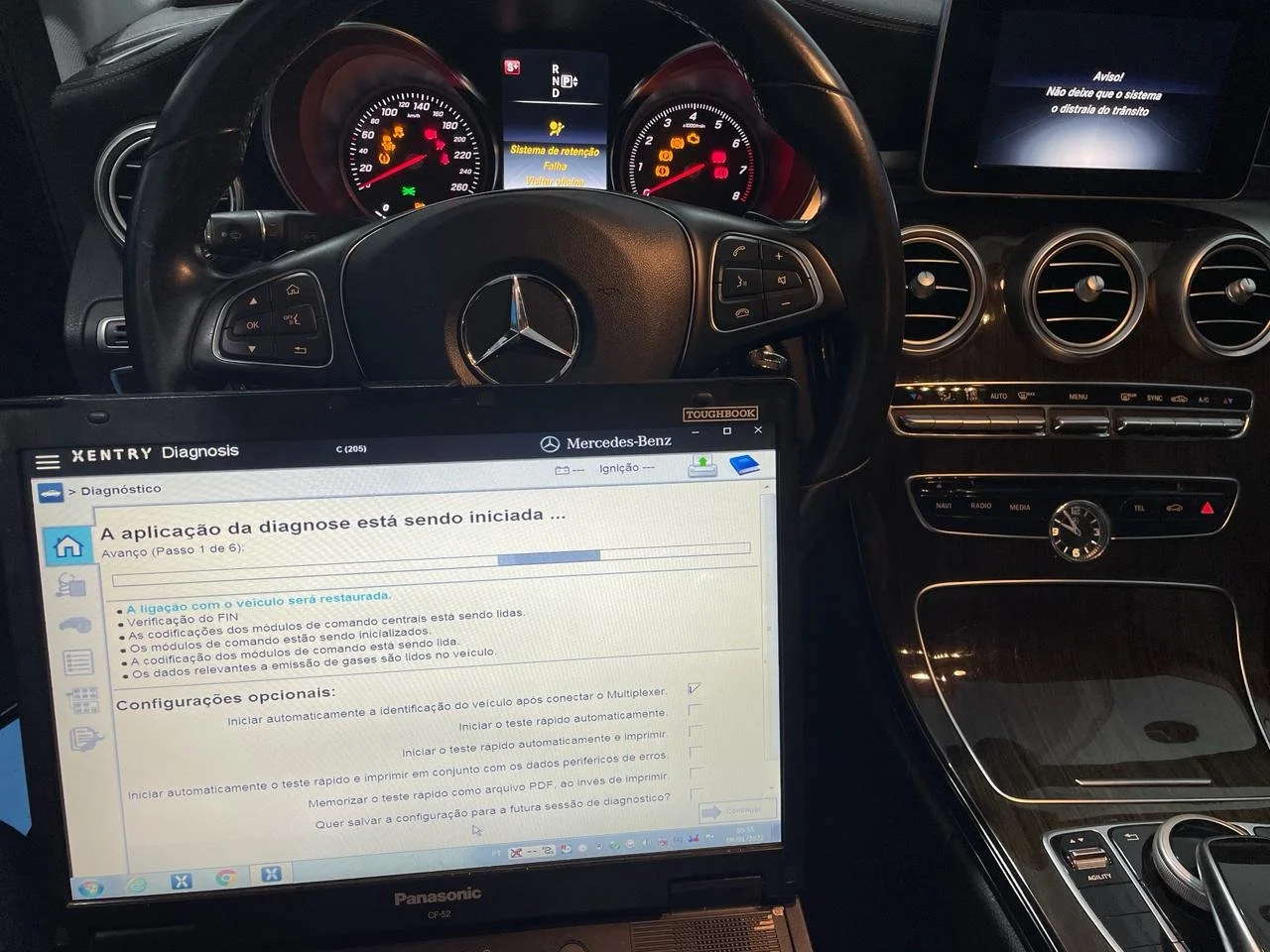 Interior de um carro Mercedes-Benz com painel digital, volante com controles, tela de diagnóstico portátil, e console com comandos de controle de clima e navegação.