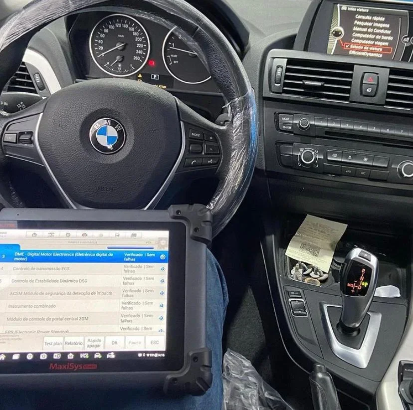 Interior de um carro BMW com painel, volante, console central e uma mesa de diagnóstico eletrônico digital ao lado, indicando procedimentos de verificação de componentes eletrônicos do veículo.