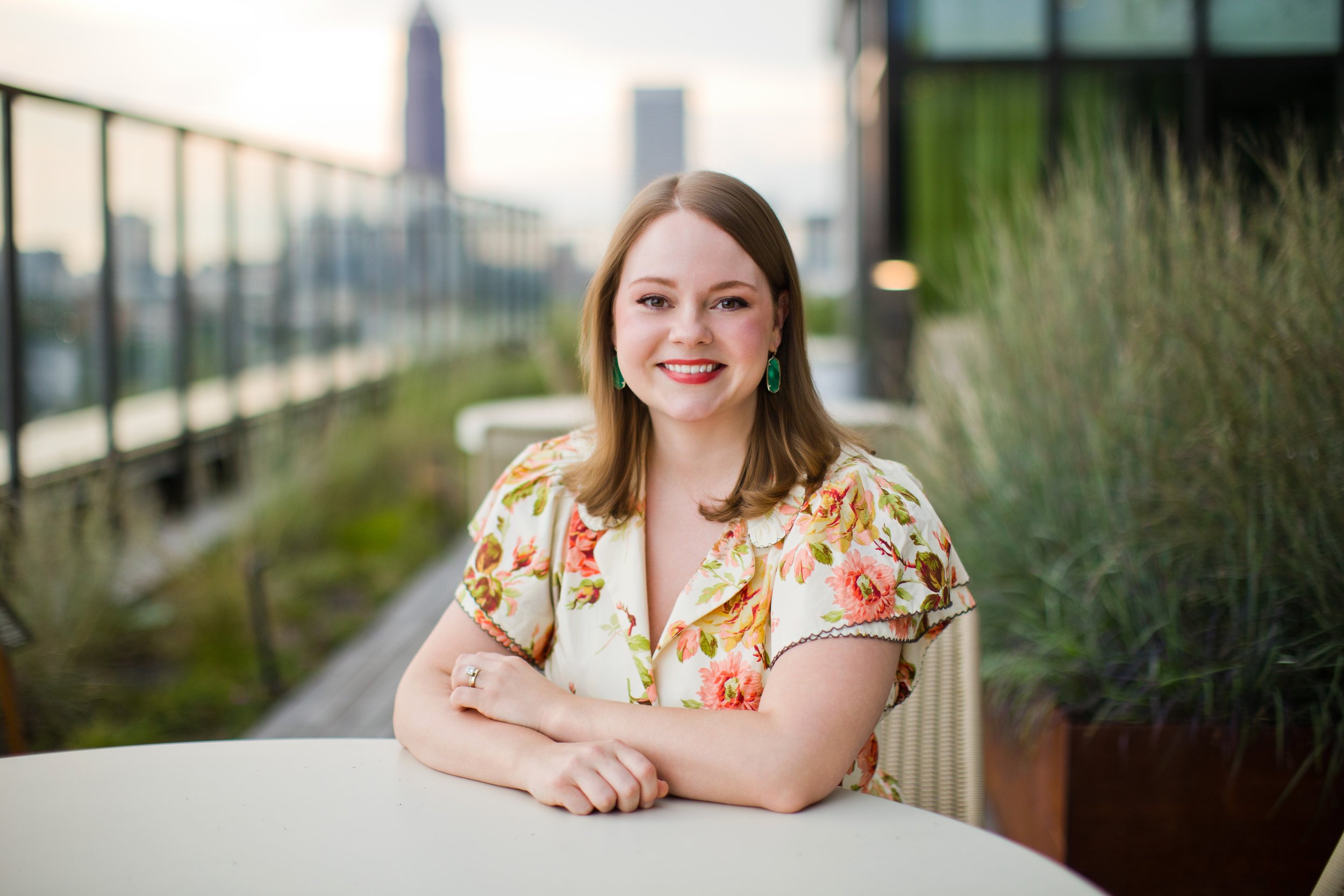 Photo of smiling woman with shoulder-length brown hair, brown eyes, wearing a cream dress with yellow, green, and rosewood florals and green earrings