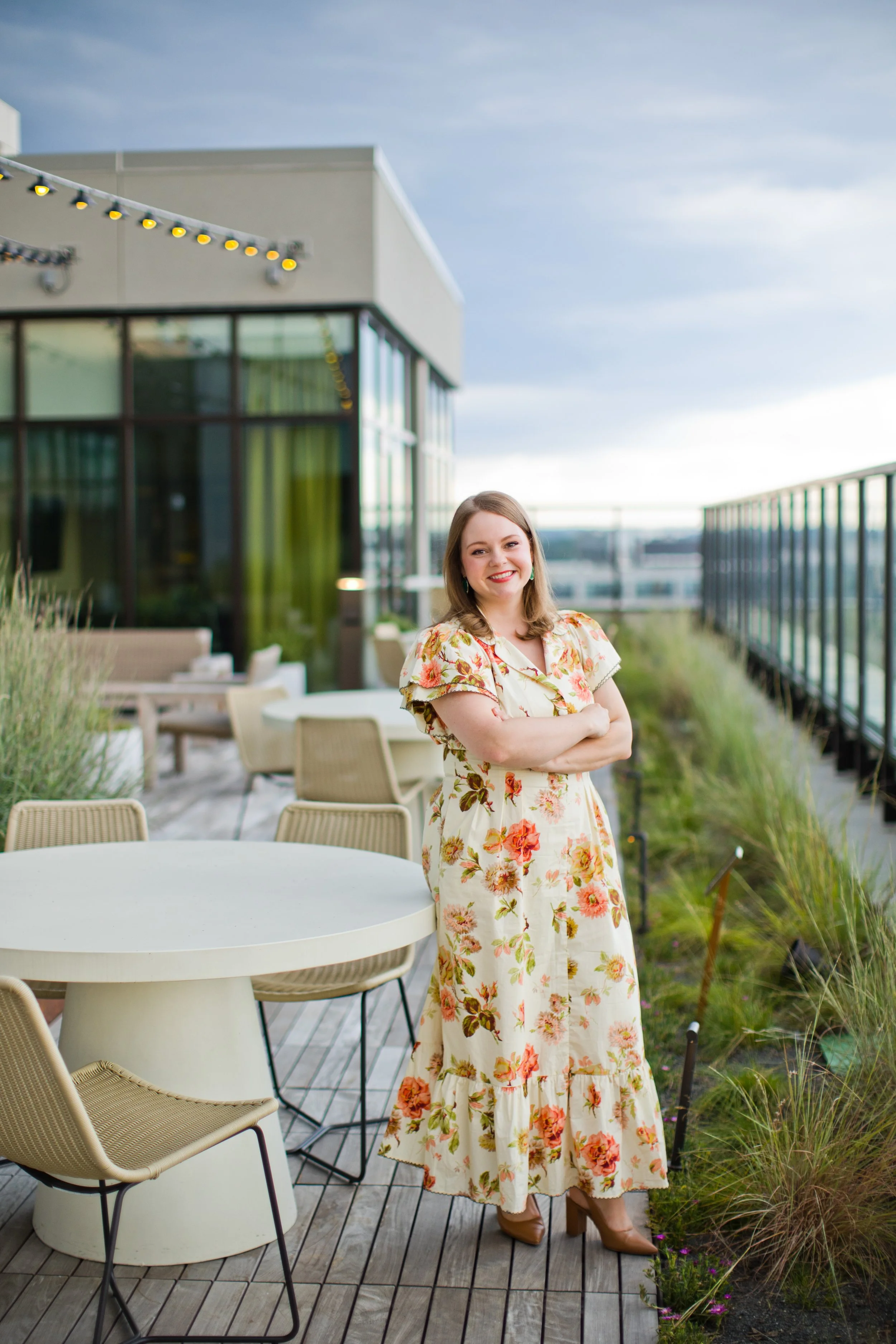 Photo of smiling woman with shoulder-length brown hair, brown eyes, wearing a cream dress with yellow, green, and rosewood florals and green earrings