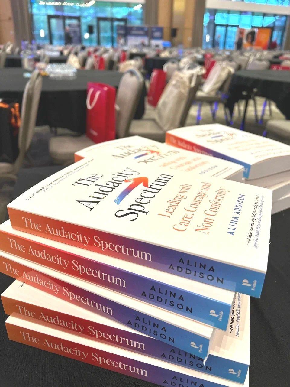 Stack of books titled 'The Audacity Spectrum' by Alina Addison on a table, with a conference or event hall with chairs and bags in the background.