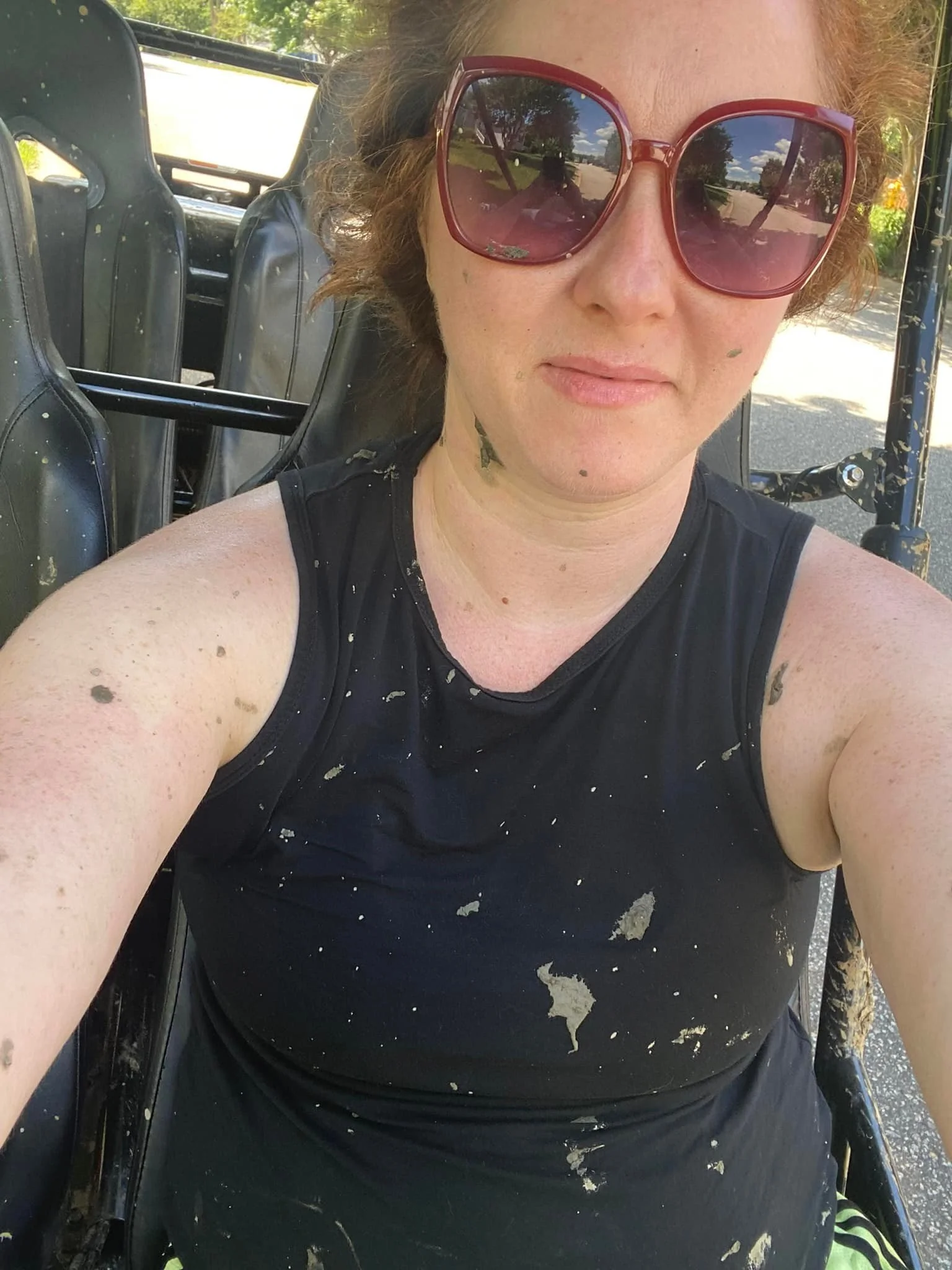 A woman wearing large red sunglasses and a black sleeveless top, sitting in a vehicle with paint splatters on her skin and clothing, outdoors on a sunny day.