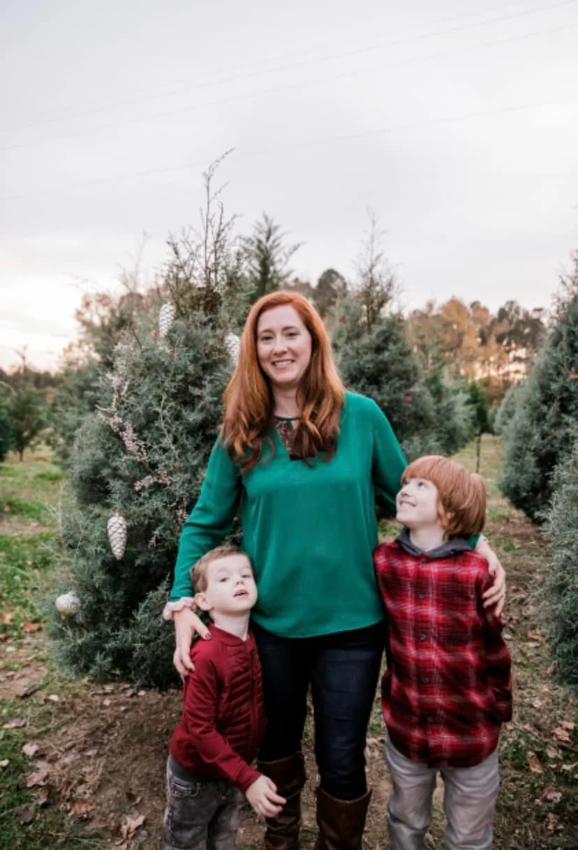 A woman with red hair standing outdoors with two young boys, surrounded by evergreen trees decorated with white pinecones, during late afternoon or early evening.