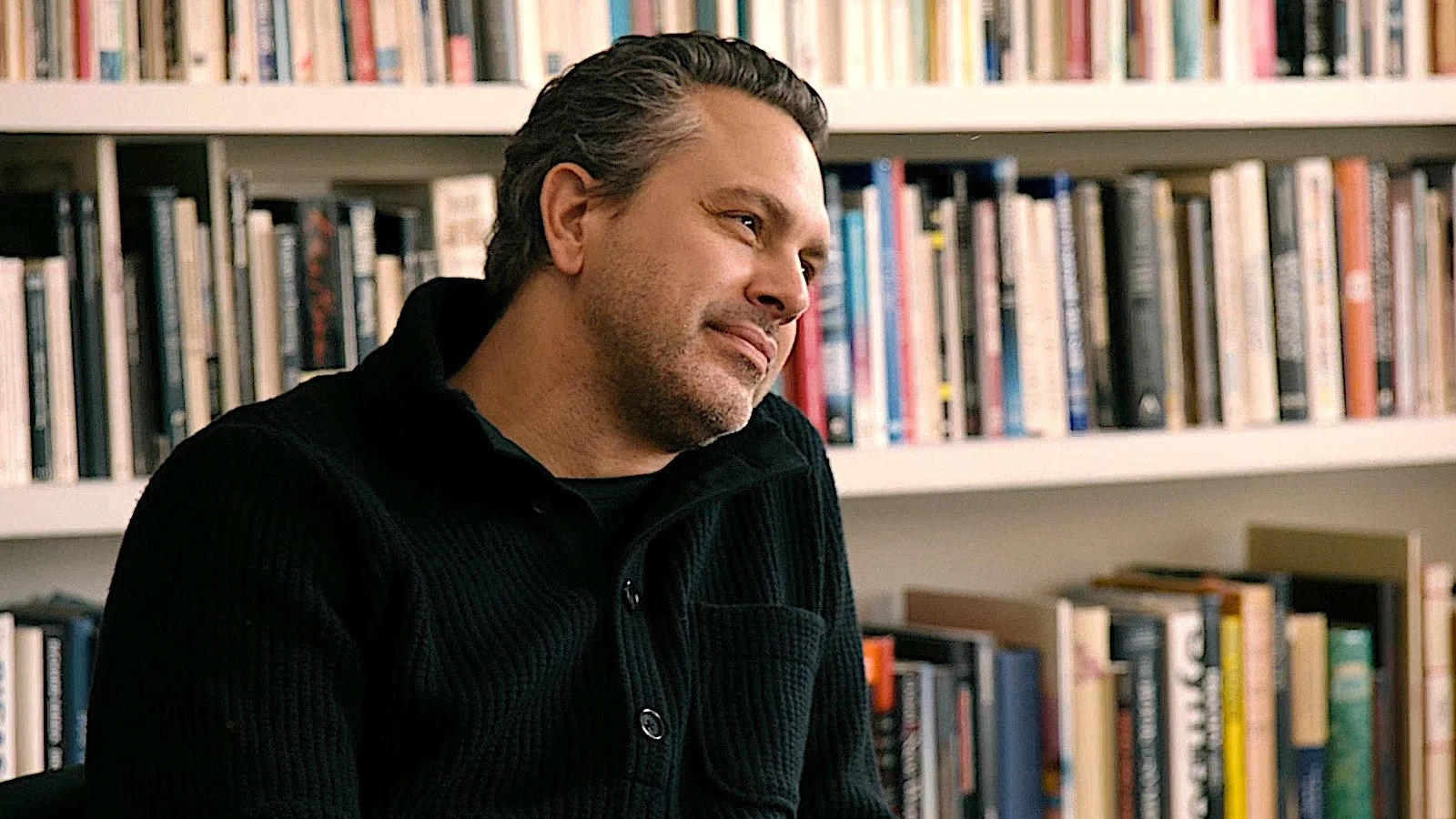 A man with dark hair and a beard sitting in front of a bookshelf filled with books, wearing a black sweater.
