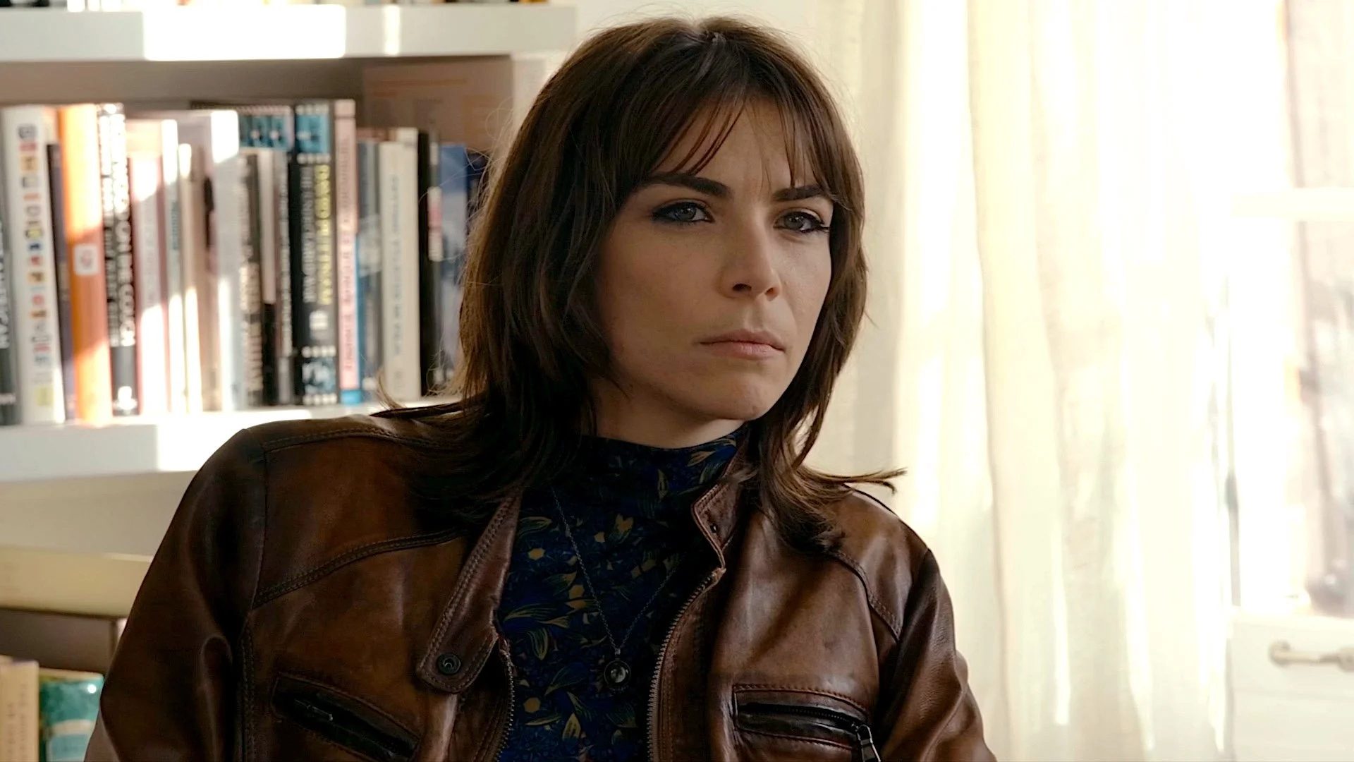 A woman with shoulder-length brown hair and blue eyes sitting indoors near a bookshelf and sheer curtains.