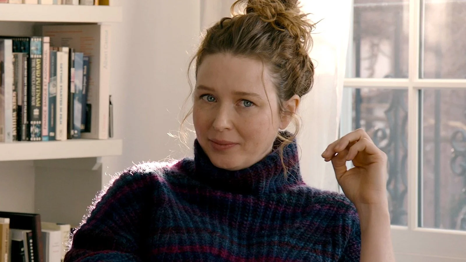 A woman with curly hair tied up, wearing a colorful sweater, sitting in a sunlit room near a window with a bookcase in the background.