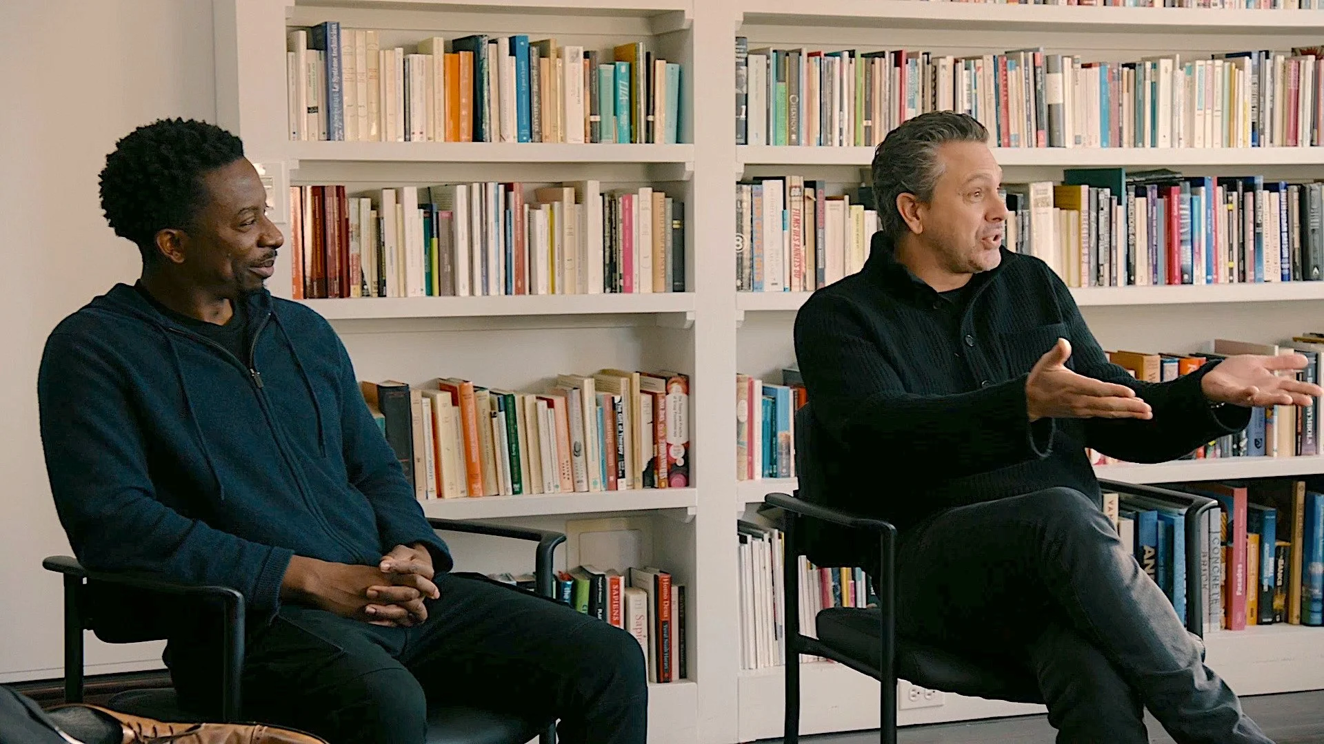 Two men sitting in chairs having a conversation in a room with white bookshelves filled with books in the background.