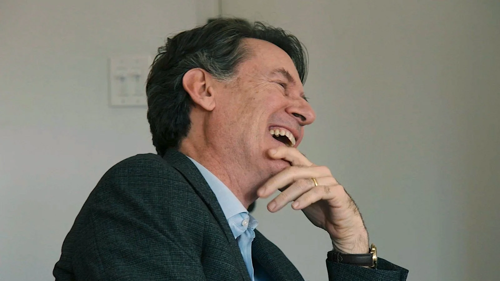 A man with dark hair, wearing a gray suit and a light blue shirt, is laughing with his eyes closed and his mouth open, holding his chin with his right hand, wearing a wedding ring and a watch.