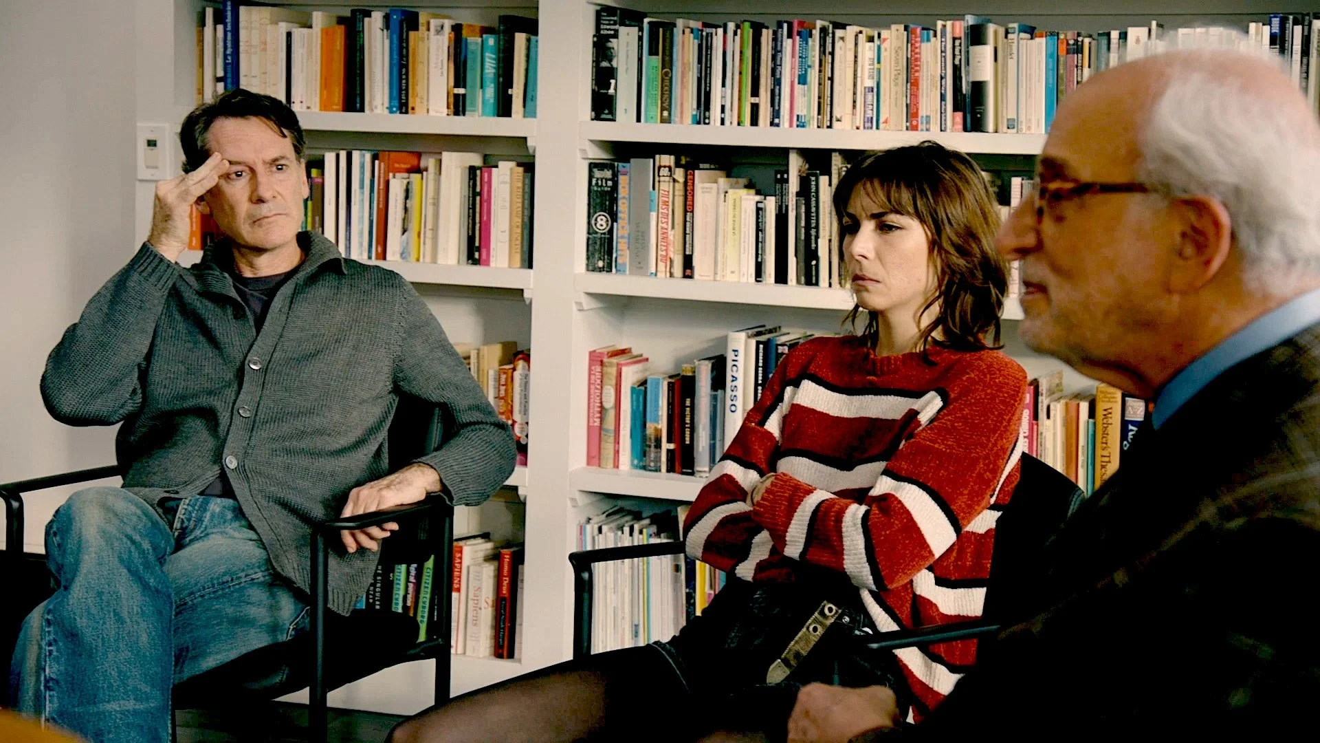 Three people sitting in a room with white bookshelves filled with books behind them, engaged in a serious conversation. One man on the left with dark hair, wearing a gray sweater, has his hand on his head. A woman in the middle with short brown hair,