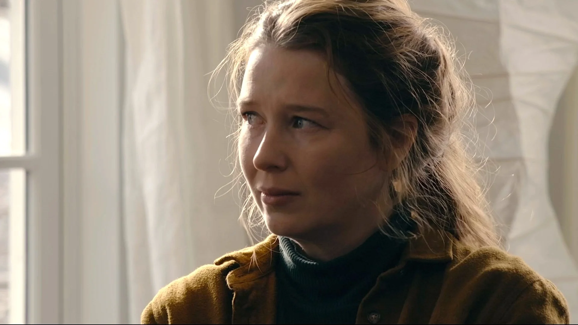 A woman with wavy brown hair and light skin gazing thoughtfully to the side, wearing a brown jacket and a black turtleneck, seated in front of a window with white curtains.