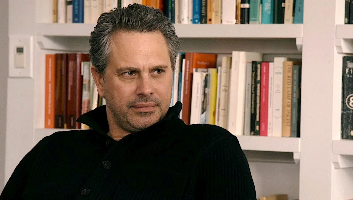 A man with gray hair and stubble, wearing a black sweater, is sitting in front of a bookshelf filled with colorful books.