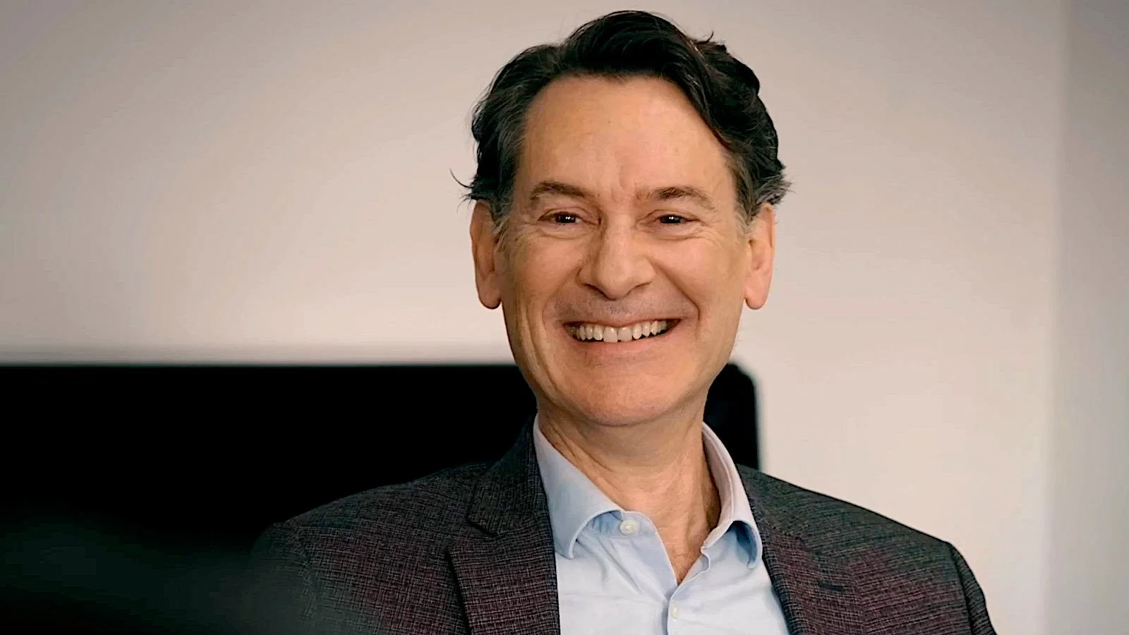 A smiling middle-aged man with dark hair, wearing a light blue dress shirt and a dark blazer, in front of a plain background.