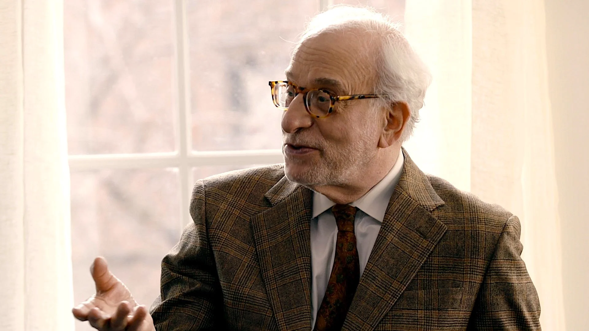 An elderly man with white hair, glasses, and a beard, wearing a brown plaid jacket and a patterned tie, gesturing with his hand while speaking near a window with light blocking curtains.