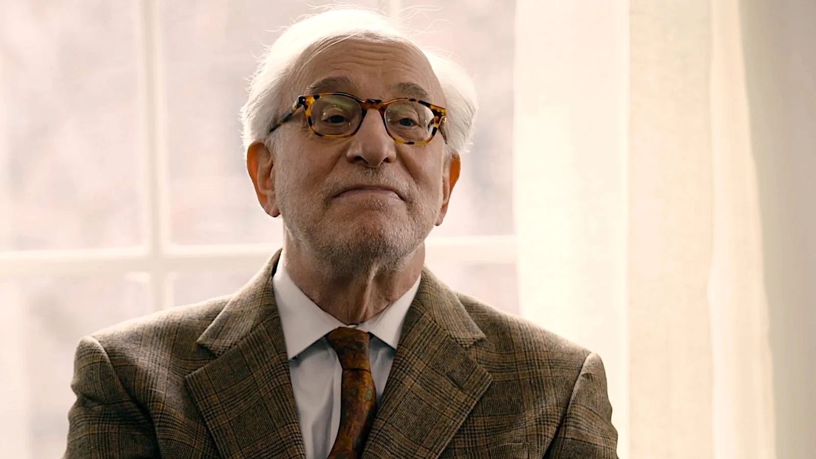 An older man with white hair and glasses, wearing a brown plaid blazer, white shirt, and brown tie, smiling softly indoors.