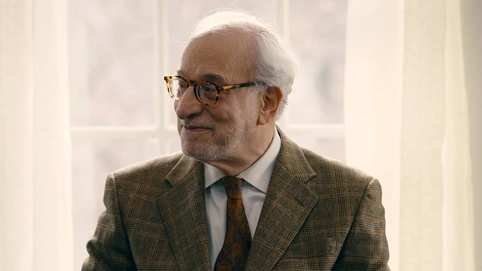 Older man with white hair, glasses, and a beard, wearing a plaid blazer and a patterned tie, sitting in front of a window with sheer curtains.