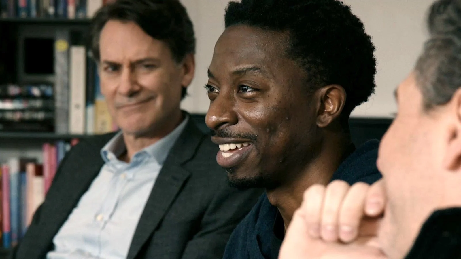 Three men are sitting together in a room, engaging in a conversation. The man in the middle is smiling, the man on the left has a smirk, and the man on the right's face is partially visible.