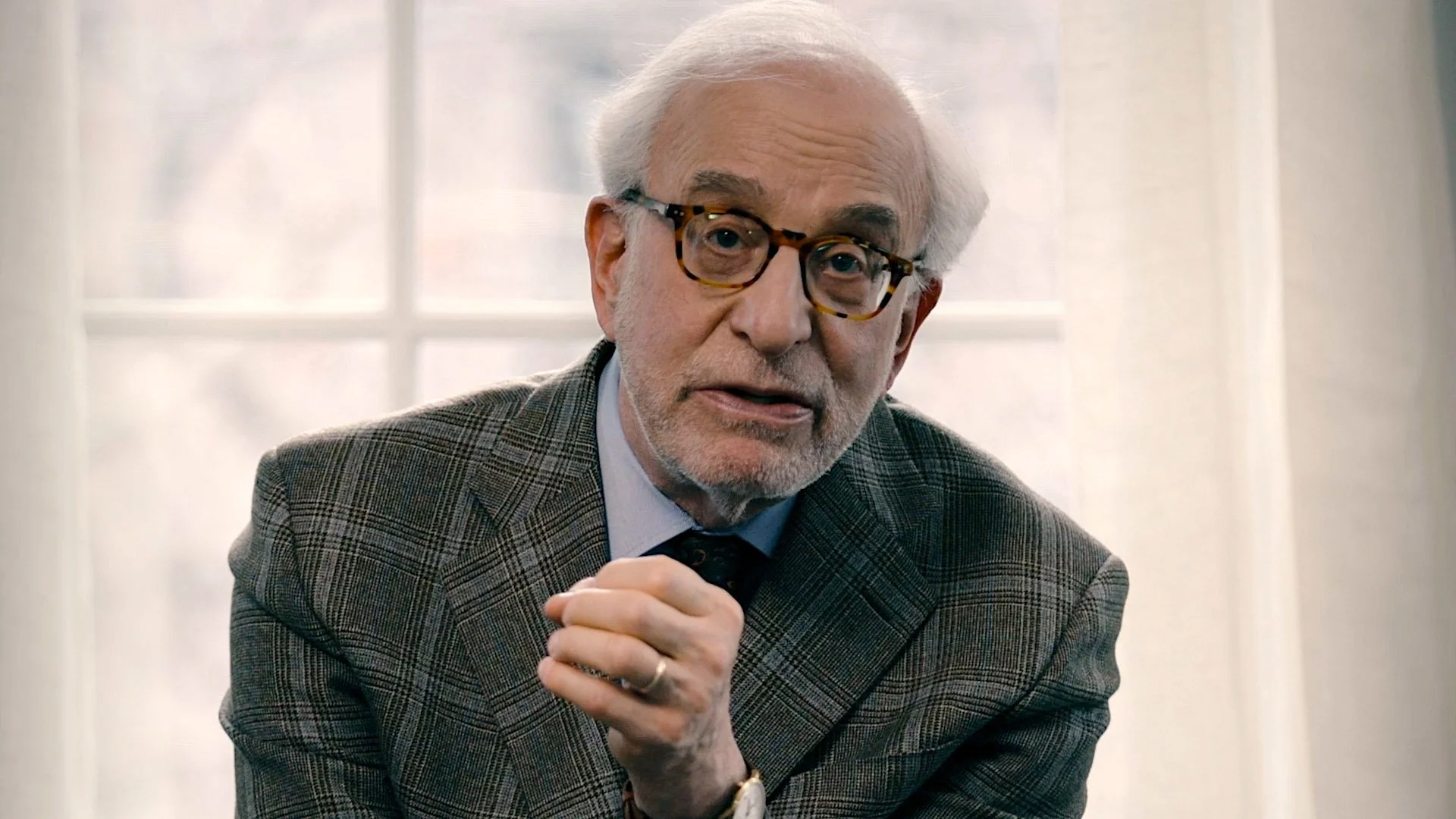 An elderly man with white hair, glasses, and a beard, wearing a checked blazer, appears to be speaking or explaining something during an interview or conversation indoors, with a window in the background.