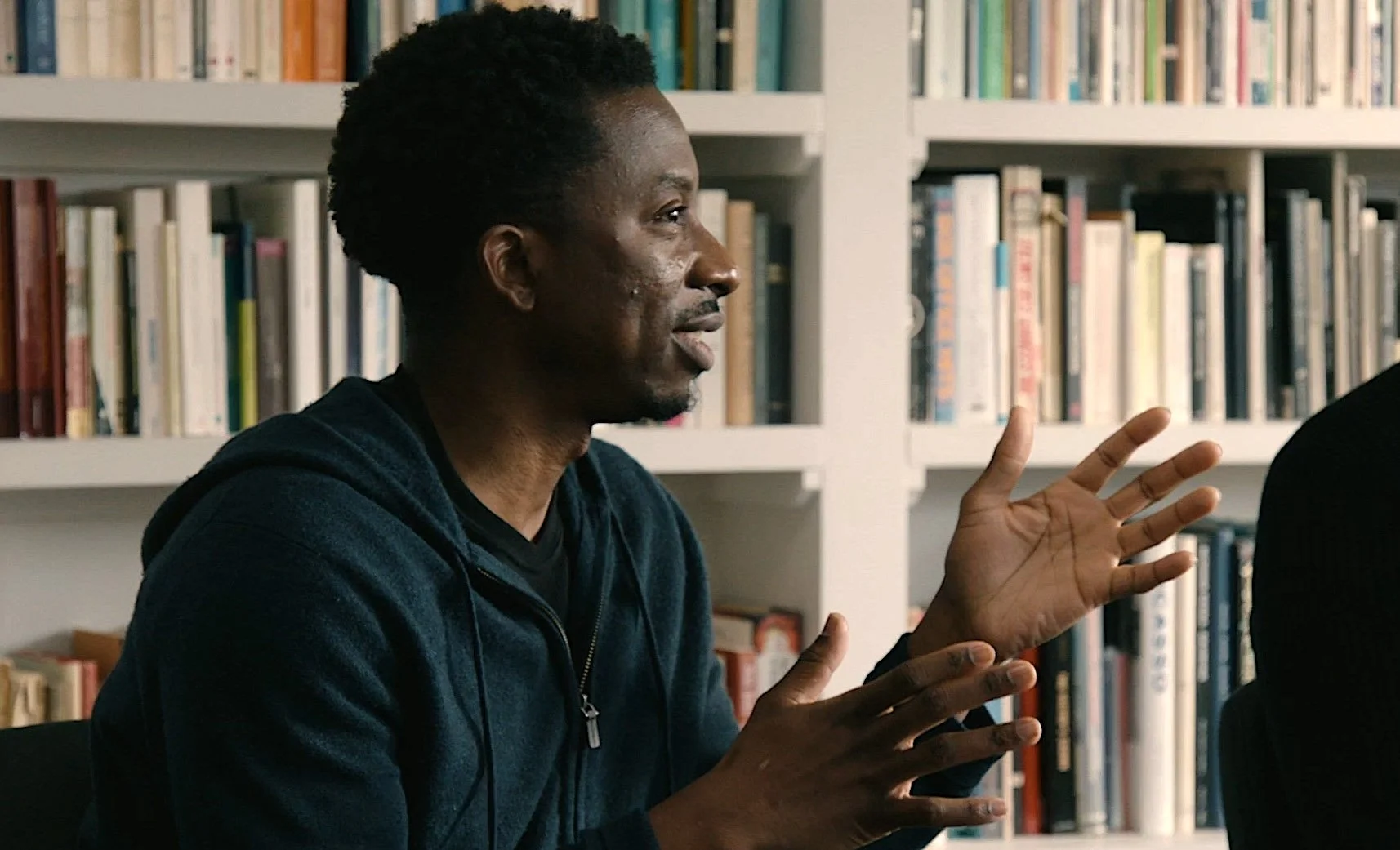 A man with short curly black hair, wearing a dark zip-up hoodie, is speaking and gesturing with his hands in front of bookshelves filled with books.