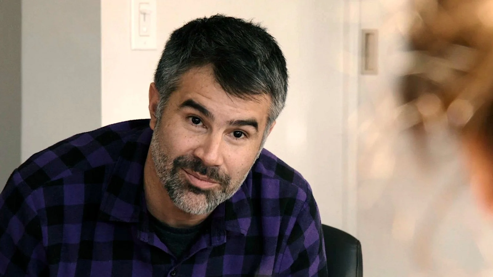 A man with short dark hair and a beard, wearing a purple and black checkered shirt, sitting indoors and looking at the camera.