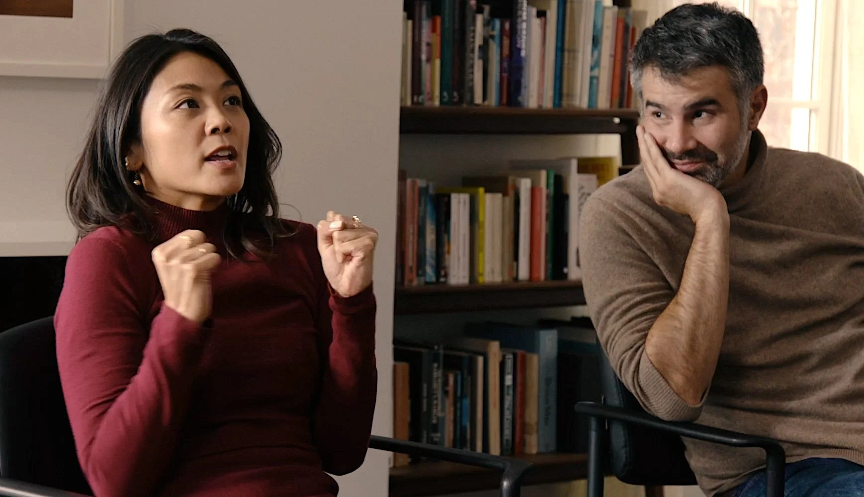 A woman with dark hair in a red long-sleeve top is speaking passionately, with her fists clenched in front of her chest. A man with salt-and-pepper hair and beard, wearing a brown sweater, is sitting with his head resting on his hand, listening and s