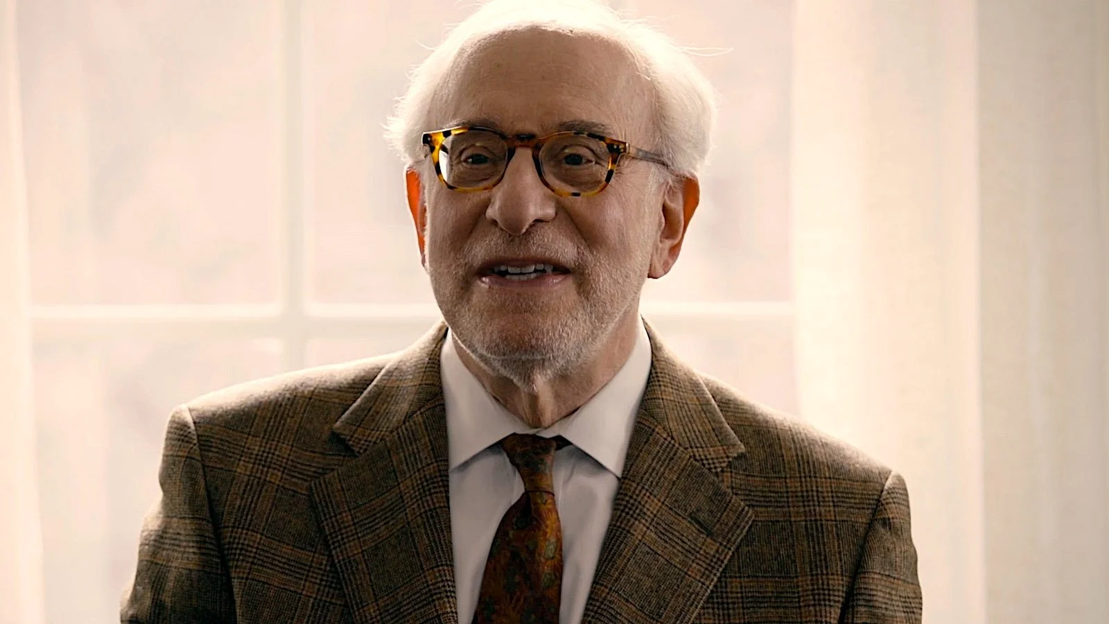 Older man with white hair, glasses, and a beard, wearing a plaid blazer, white shirt, and patterned tie, speaking in front of a window with sheer curtains.