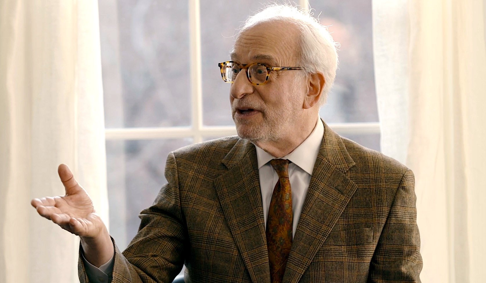 Elderly man with glasses wearing a plaid blazer and tie speaking and gesturing with his left hand in front of a window with white curtains.