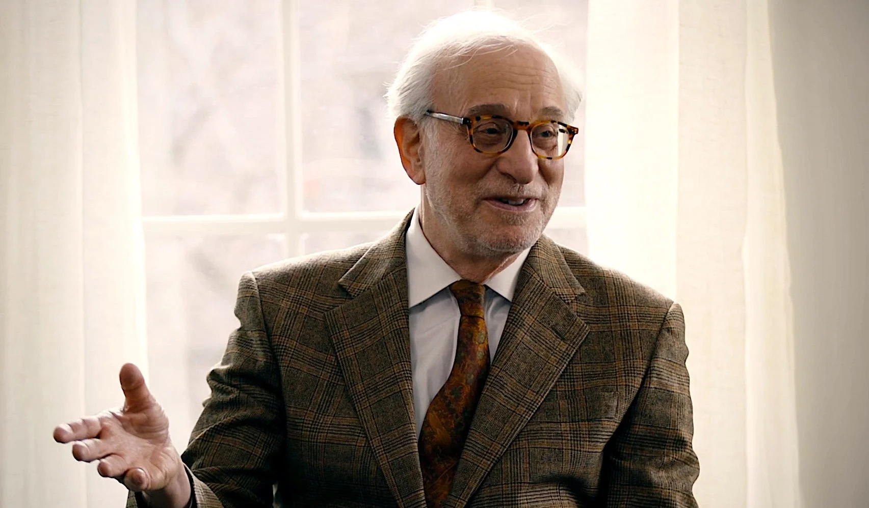 Older man with white hair and glasses, wearing a brown plaid suit and a patterned tie, smiling and gesturing with his hand in a brightly lit room with curtains behind him.