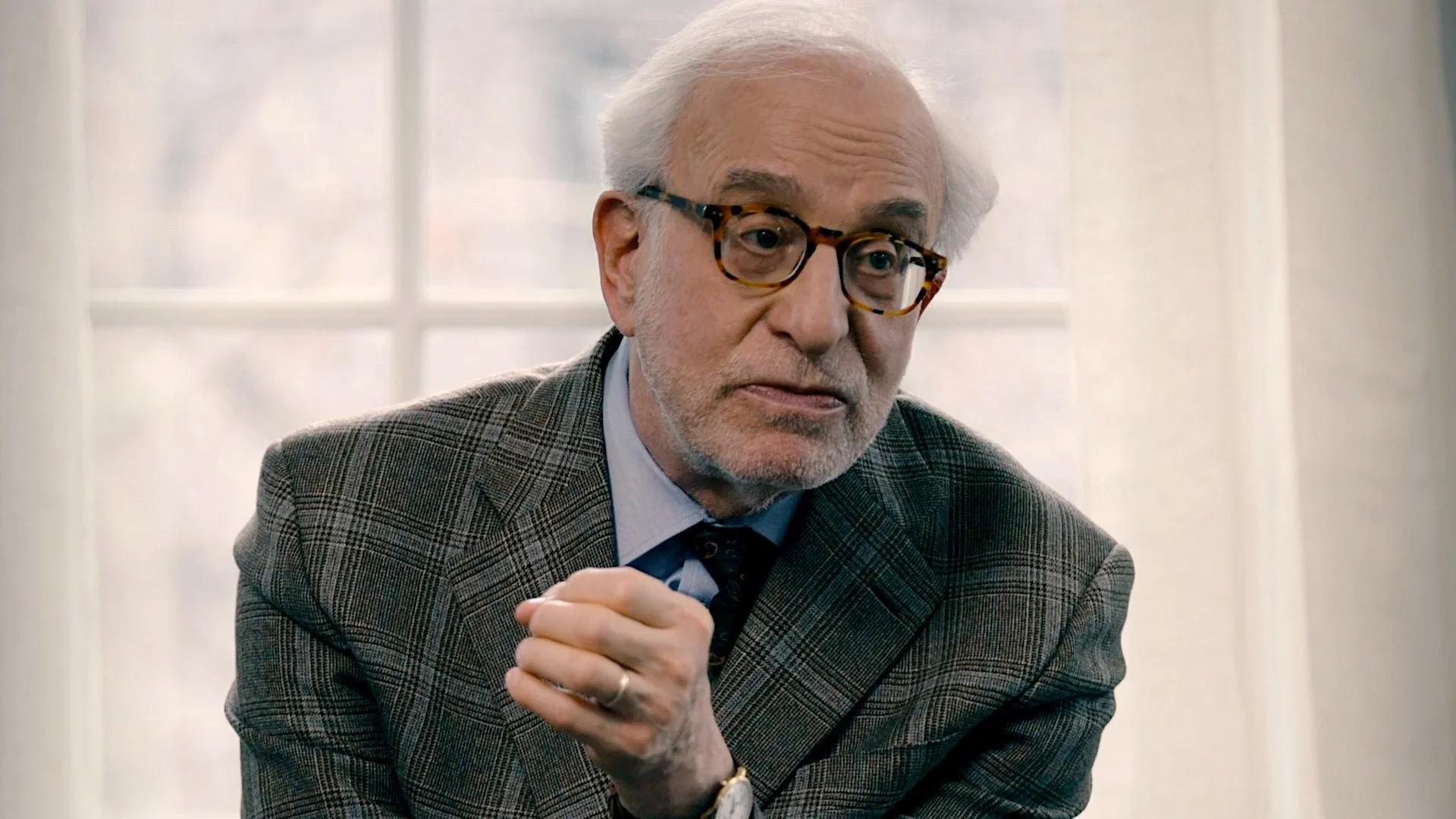 An elderly man with white hair and beard, wearing glasses and a plaid jacket, sitting indoors near a window.