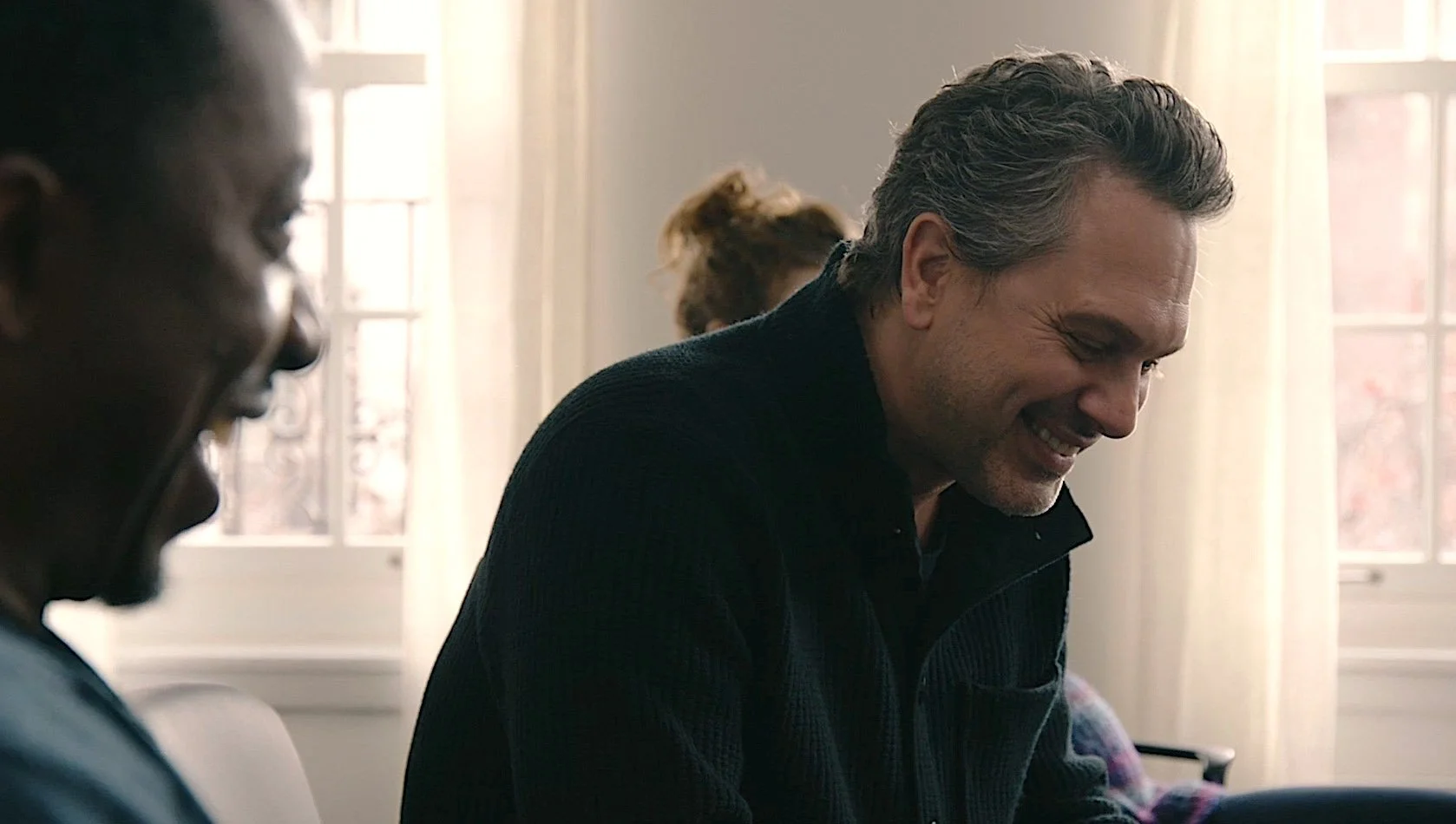 Two people smiling and sitting together indoors near a window, with sunlight coming through.