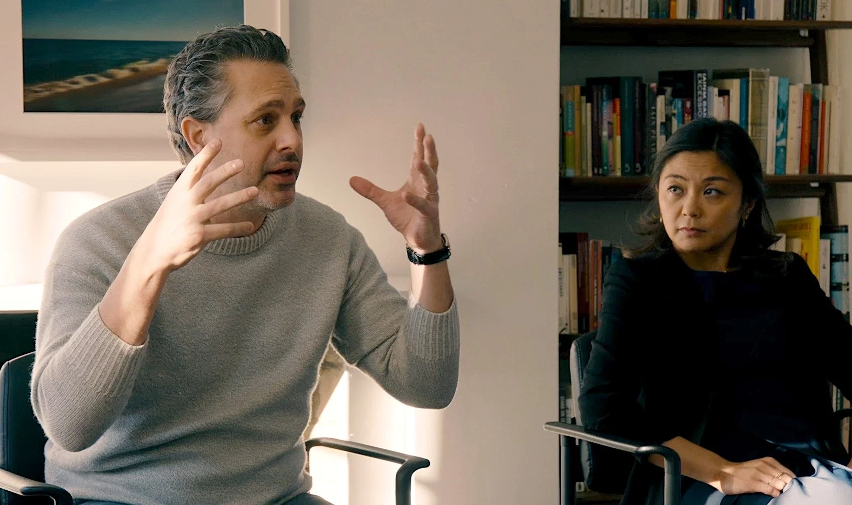 A man in a beige sweater gesturing with his hands while speaking, seated next to a woman in a black top listening in a room with bookshelves.