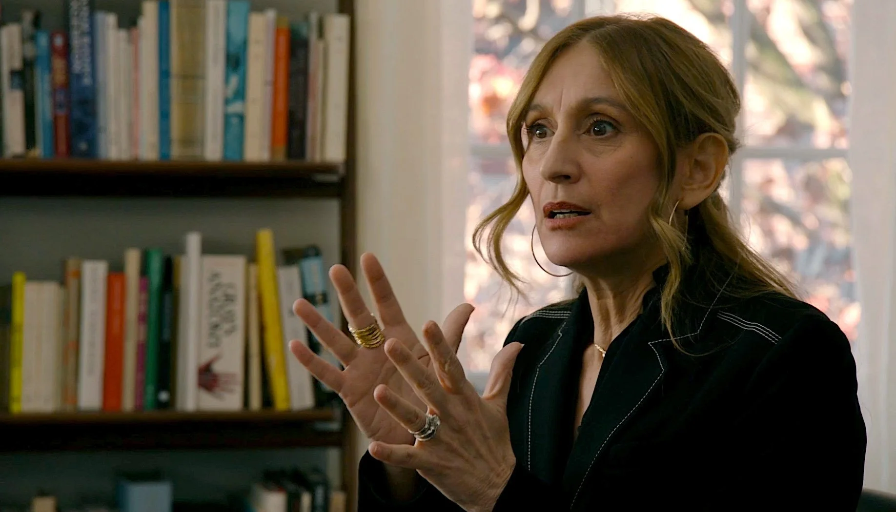A woman with reddish-brown hair wearing a black blouse is speaking and gesturing with her hands in front of a bookshelf with colorful books and a window with trees outside.