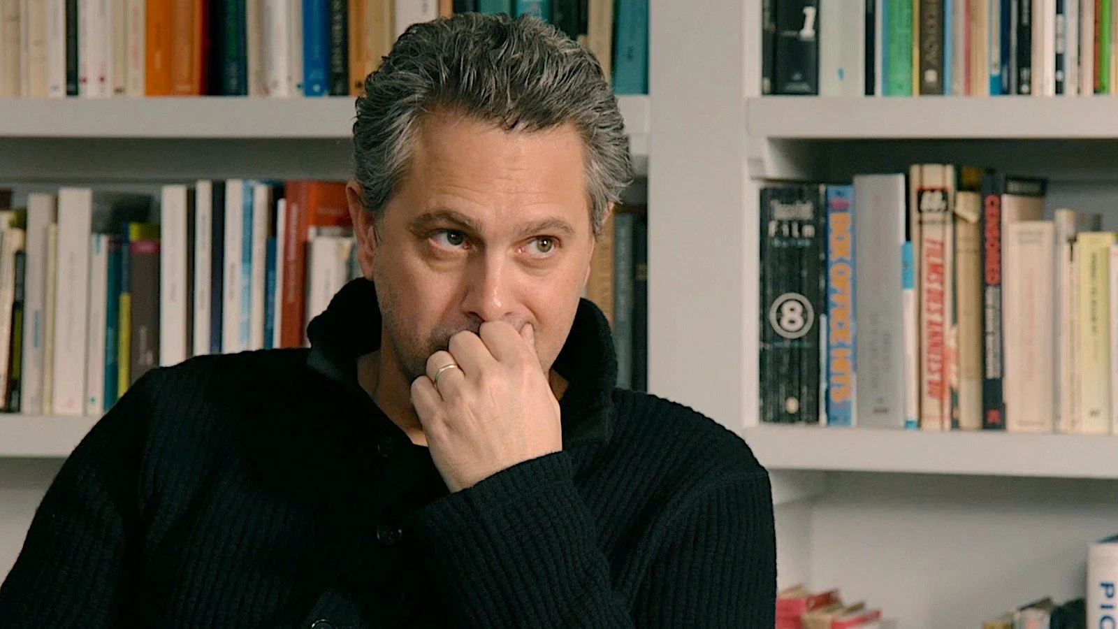 A man with gray hair and a black sweater sitting in front of a bookshelf, resting his chin on his hand, appearing thoughtful.