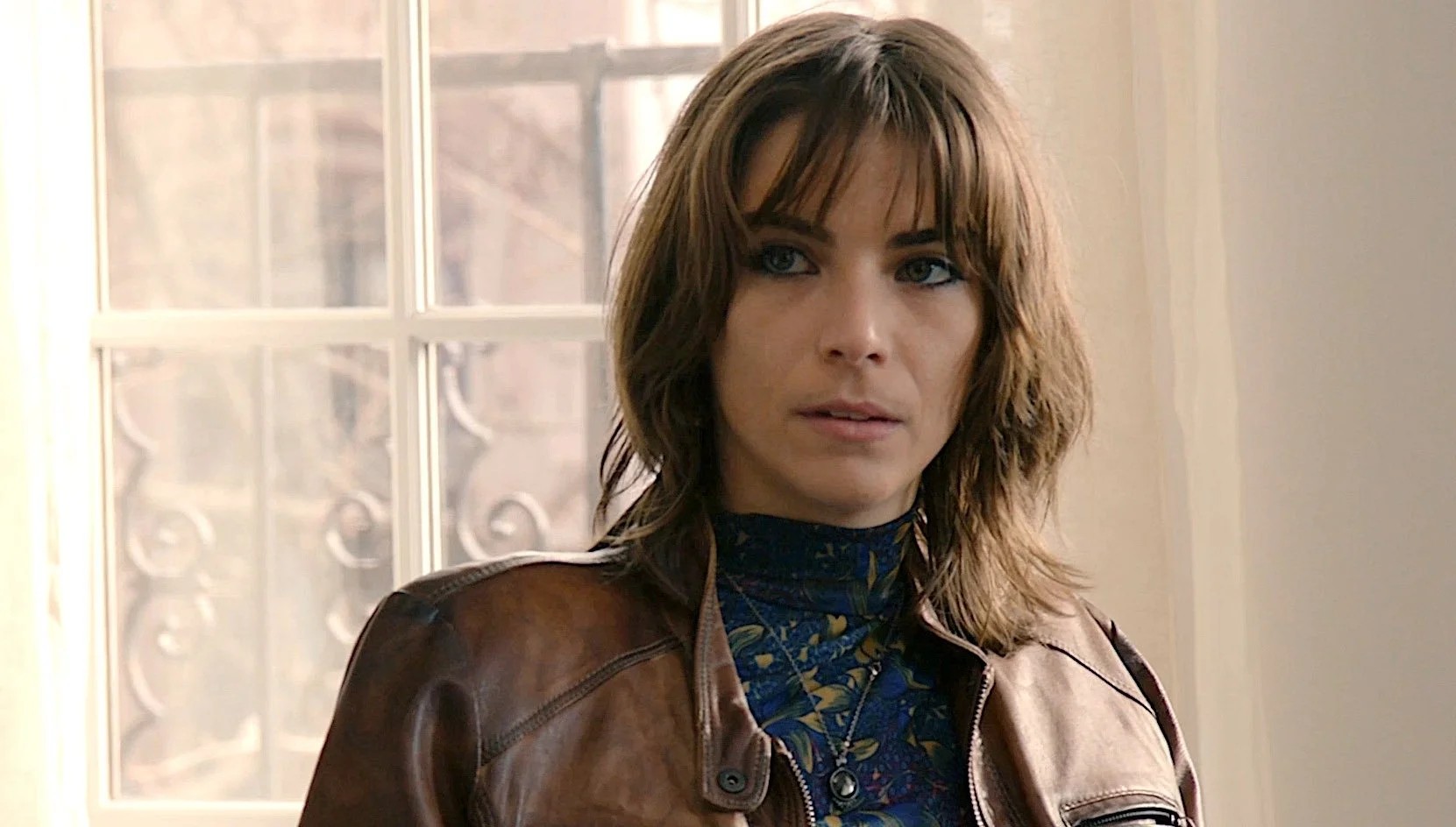 A woman with shoulder-length brown hair wearing a leather jacket and patterned turtleneck. She is standing indoors near a window.