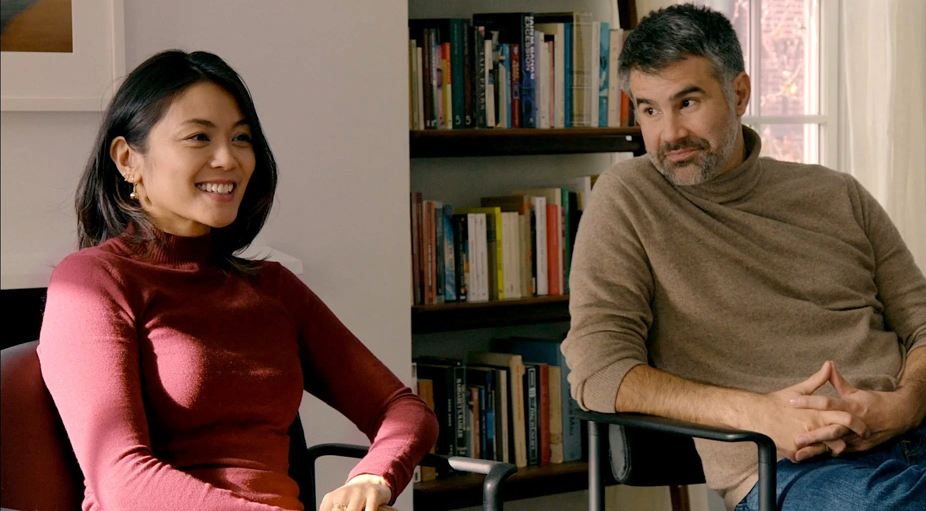 A woman with black hair, wearing a red top, is smiling and sitting next to a man with short gray hair and beard, wearing a beige turtleneck, who is attentively listening. They are seated in a room with a bookshelf filled with books and a window in th