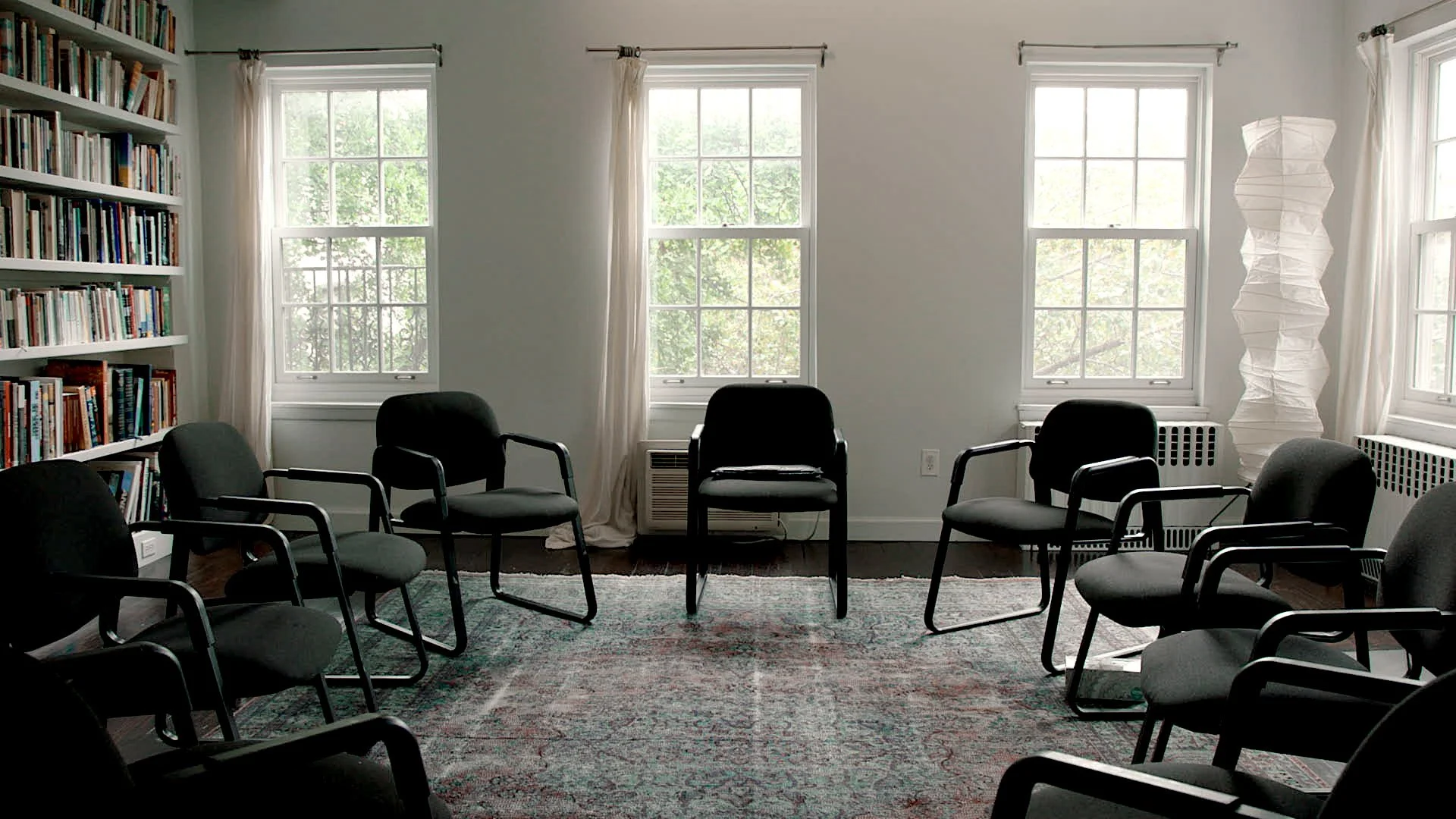 A conference or meeting room with six black chairs arranged in a circle, three large windows with white curtains, a tall paper floor lamp, a bookshelf filled with books, an area rug, and a white wall with an air conditioner and radiator.