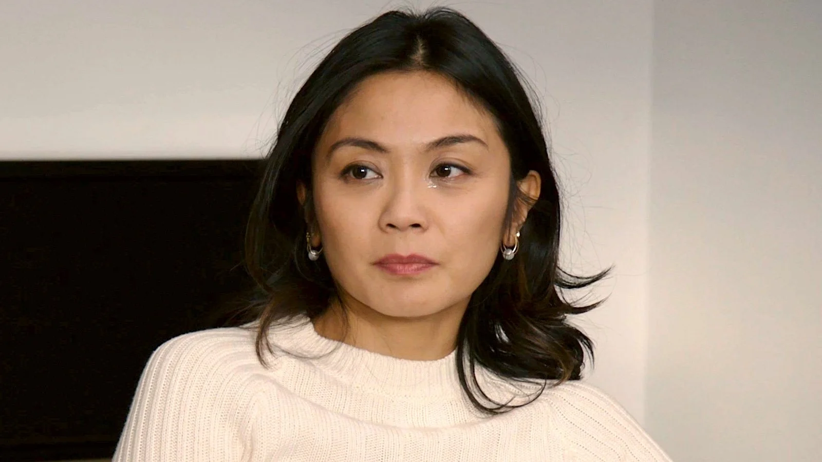A woman with shoulder-length dark hair, wearing a white sweater, sitting indoors, looking to her left with a neutral expression.