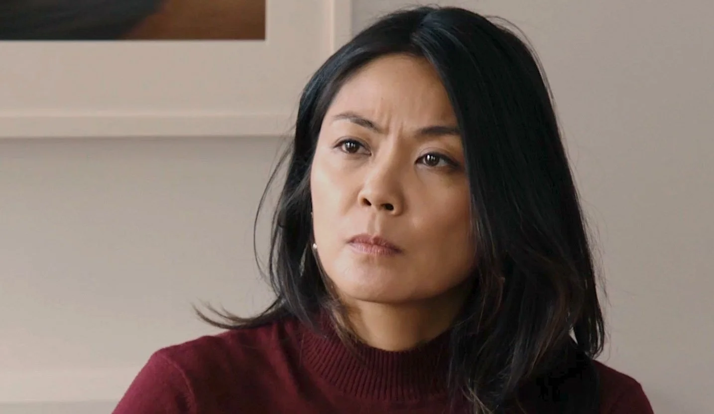 A woman with shoulder-length black hair and a serious expression, wearing a maroon sweater, sitting indoors.
