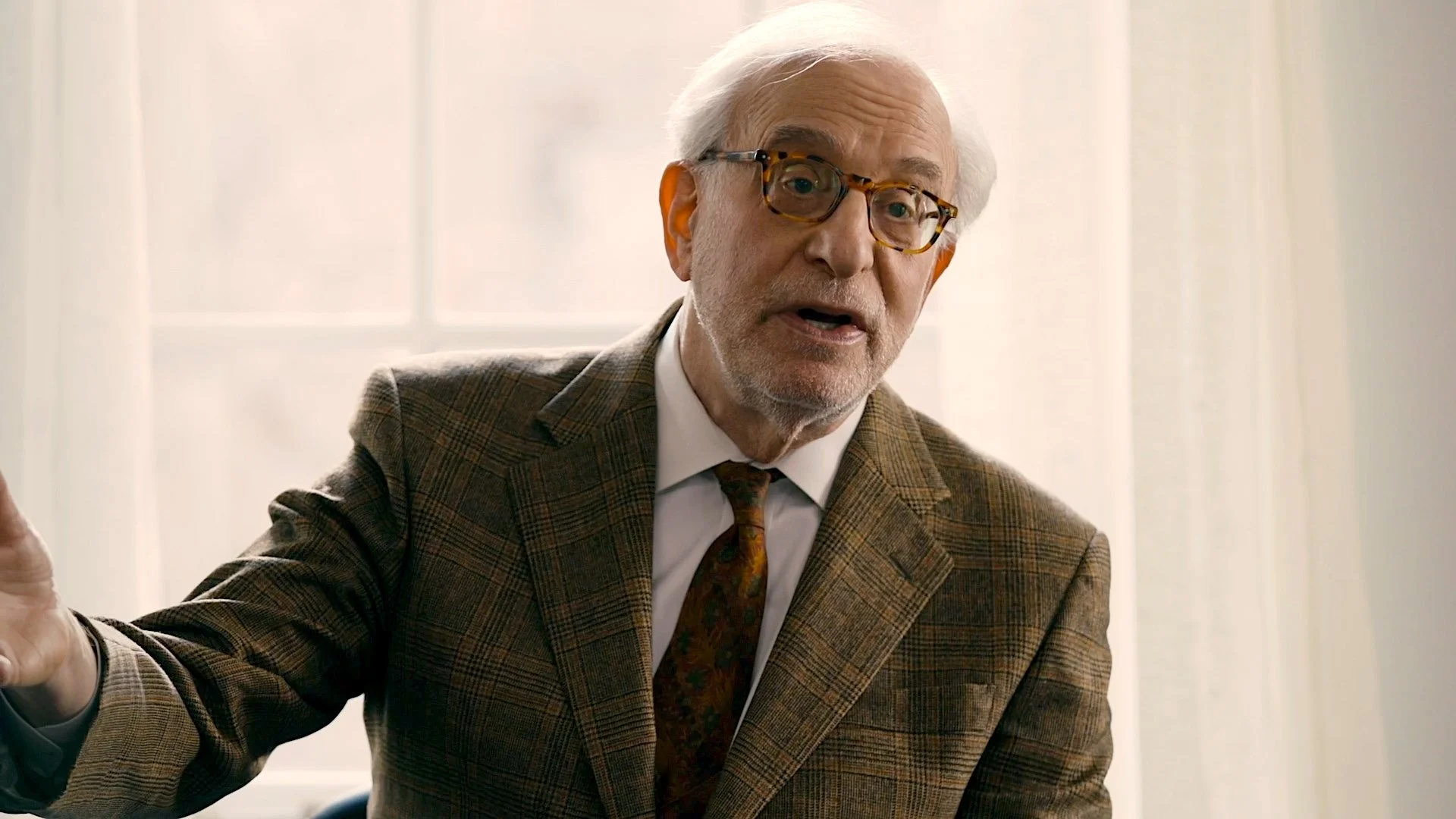 An elderly man with white hair, glasses, and a beard, wearing a brown plaid suit, white shirt, and patterned tie, appears to be speaking or explaining something in a well-lit room.