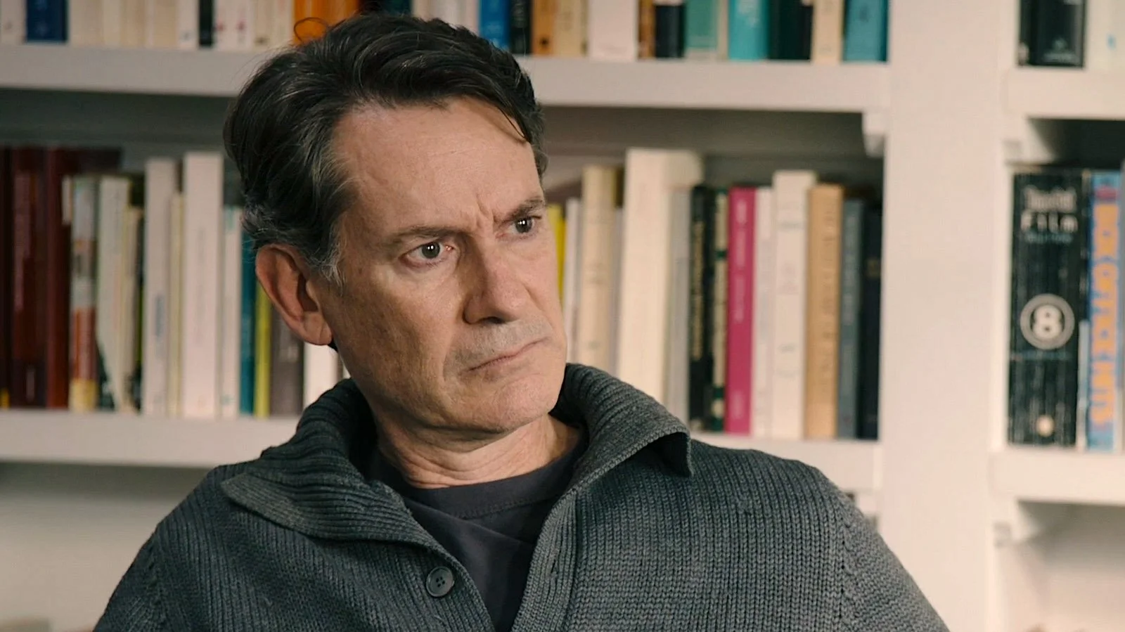 A man with dark hair and a serious expression, wearing a gray sweater, standing in front of a bookshelf filled with books.