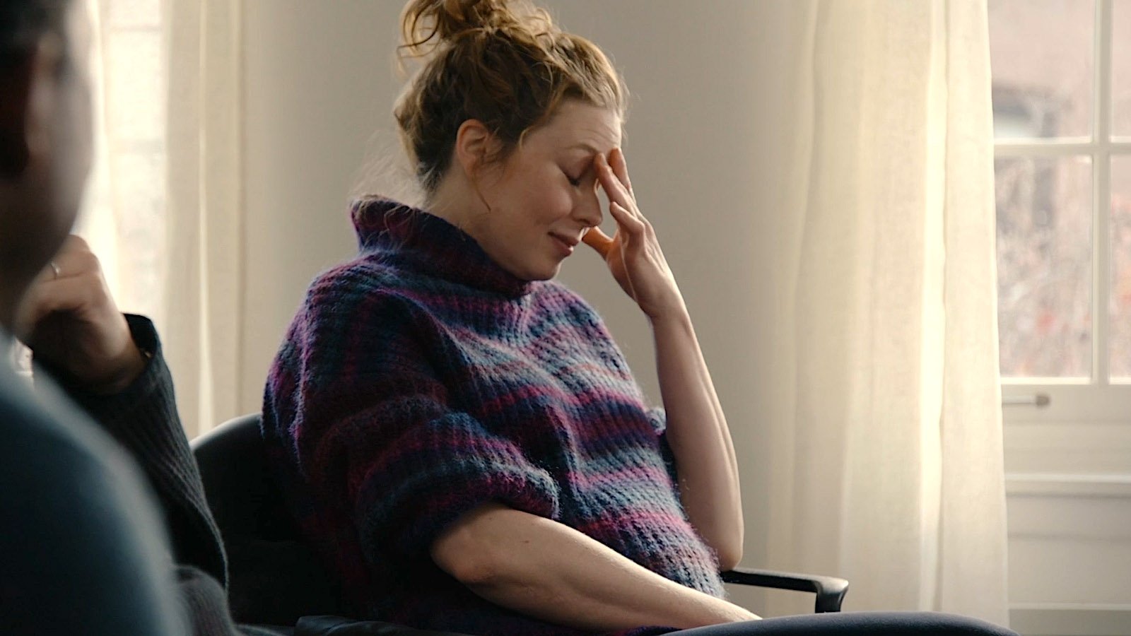 A woman with red hair in a bun sitting with her eyes closed, holding her forehead in a distressed gesture inside a room with light-colored curtains and a window in the background.
