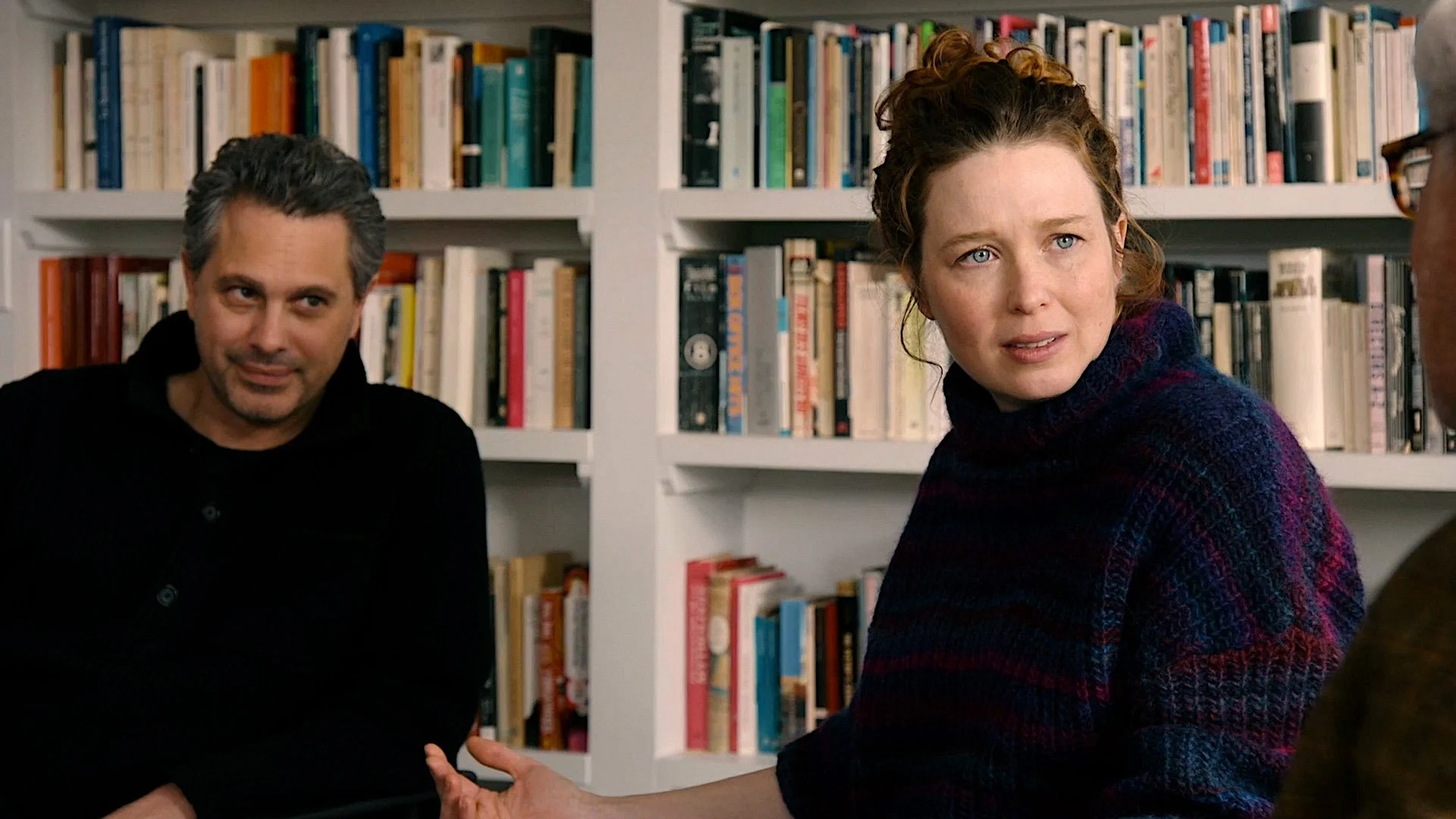 Three people are having a conversation in front of a bookshelf filled with books. The woman in the center looks concerned. The man on the left is smiling slightly, and the person on the right is wearing glasses.