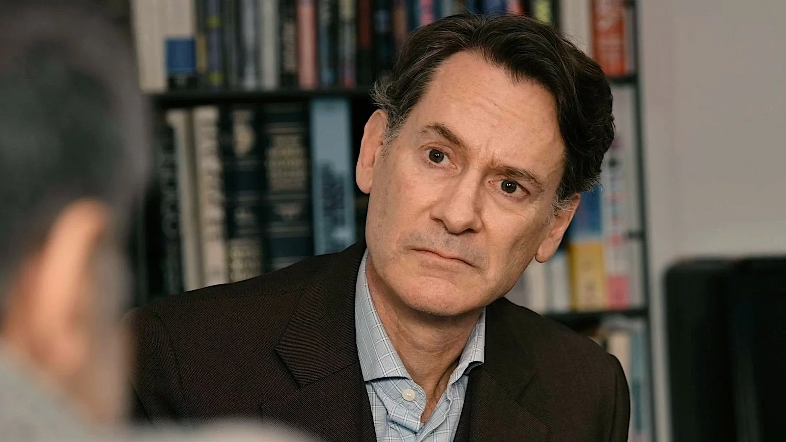 A middle-aged man with dark hair and wearing a dark suit jacket over a light button-up shirt, looking concerned or serious, sitting near a bookshelf filled with books.