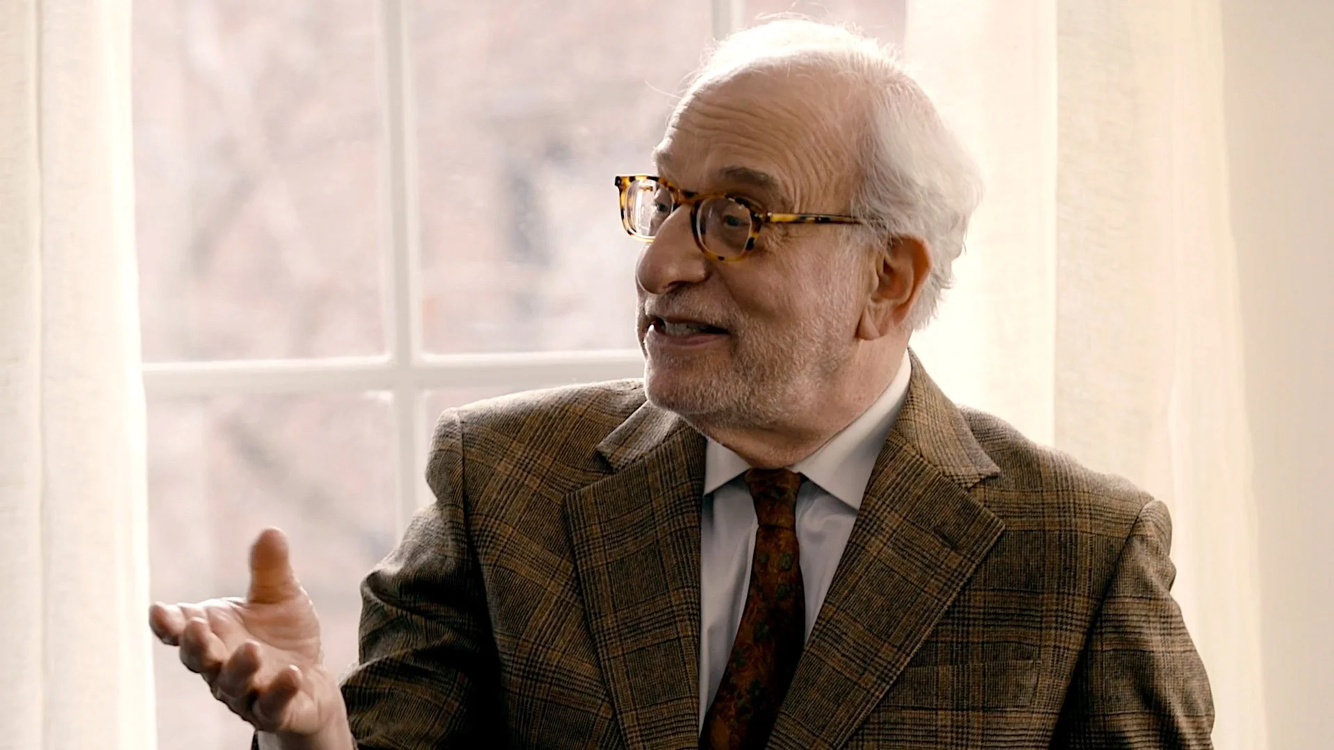 An elderly man with white hair, glasses, wearing a brown plaid suit and a dark patterned tie, is speaking and gesturing with his right hand in front of a window with white curtains.