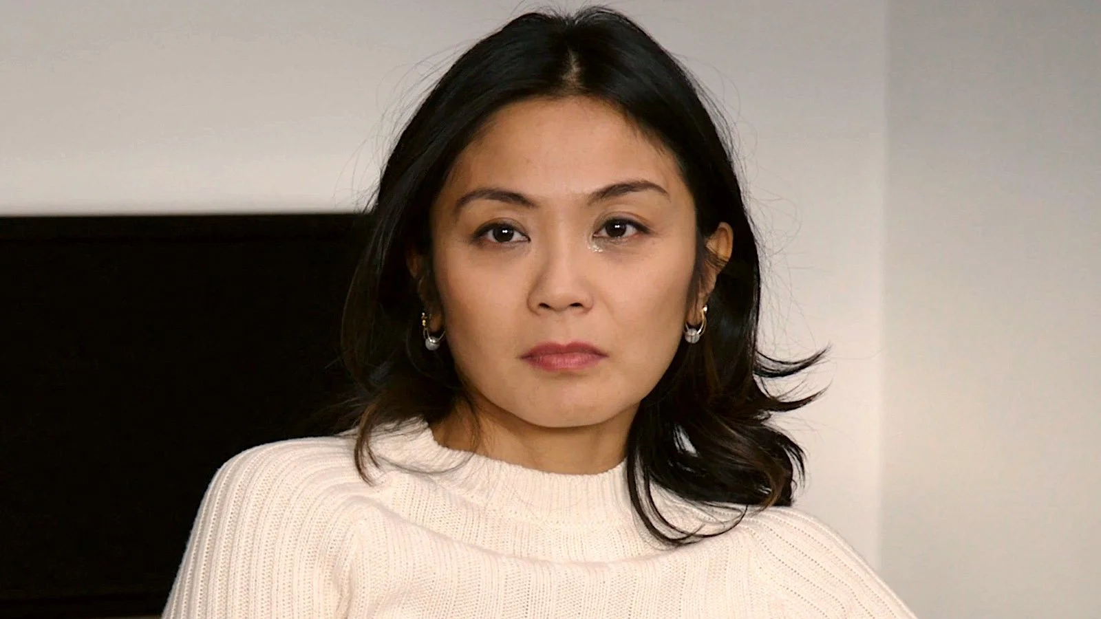 A woman with dark hair, wearing a cream-colored sweater and earrings, sitting in front of a plain background.