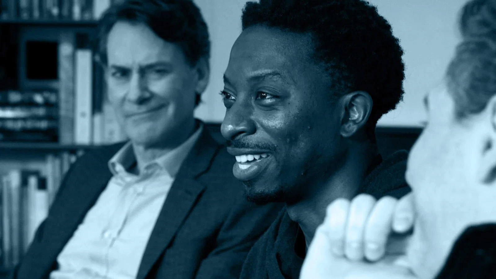 Three people engaged in a conversation in a professional setting, with a bookshelf in the background, during a meeting or discussion.