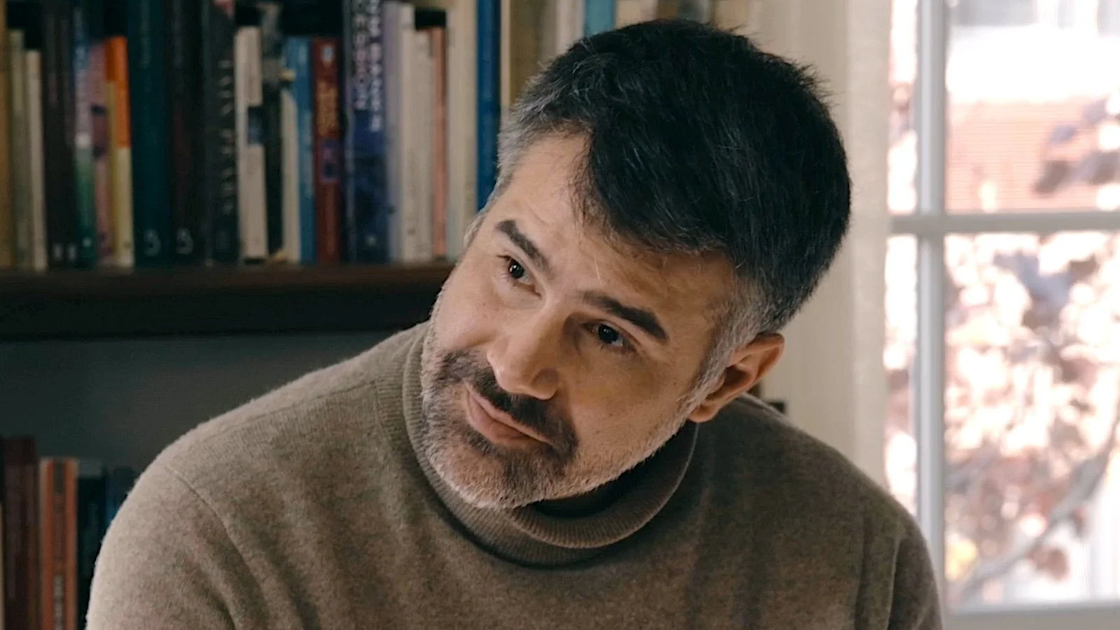 A man with dark hair and a beard sits in a room with a bookshelf behind him and a window to his side.