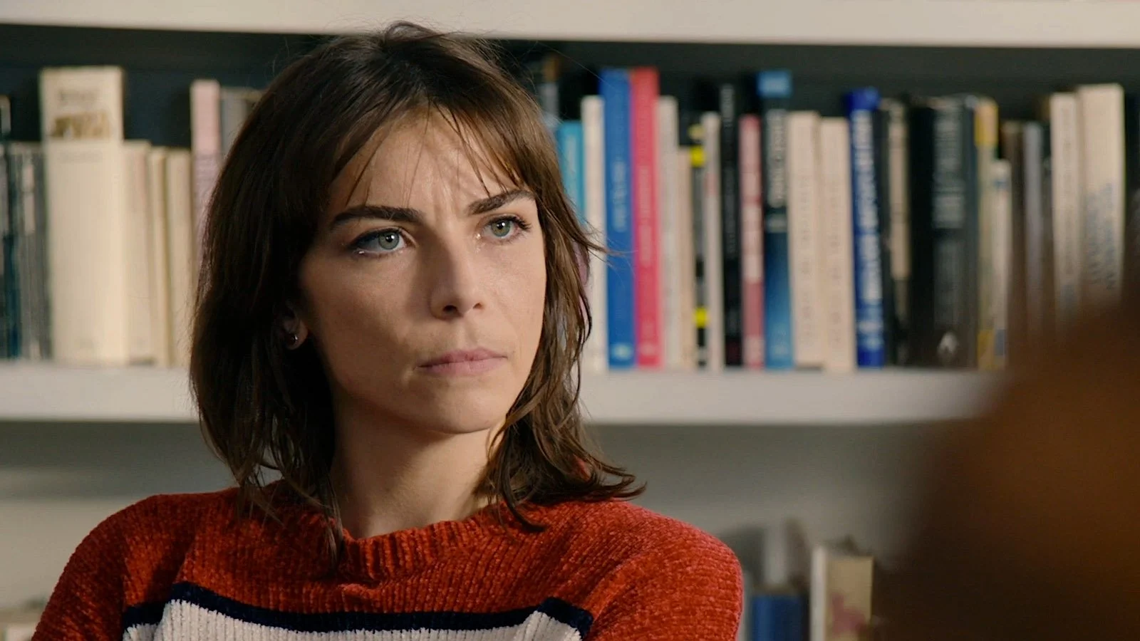 A woman with shoulder-length brown hair and blue eyes, wearing a red sweater with white and black stripes, sitting in front of a bookshelf filled with books.